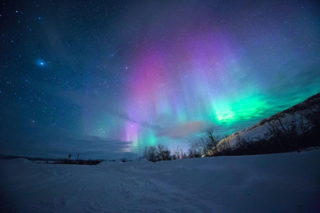 Un ciel nocturne spectaculaire rempli d'étoiles avec une aurore boréale aux teintes roses, violettes et vertes au-dessus d'un paysage enneigé.