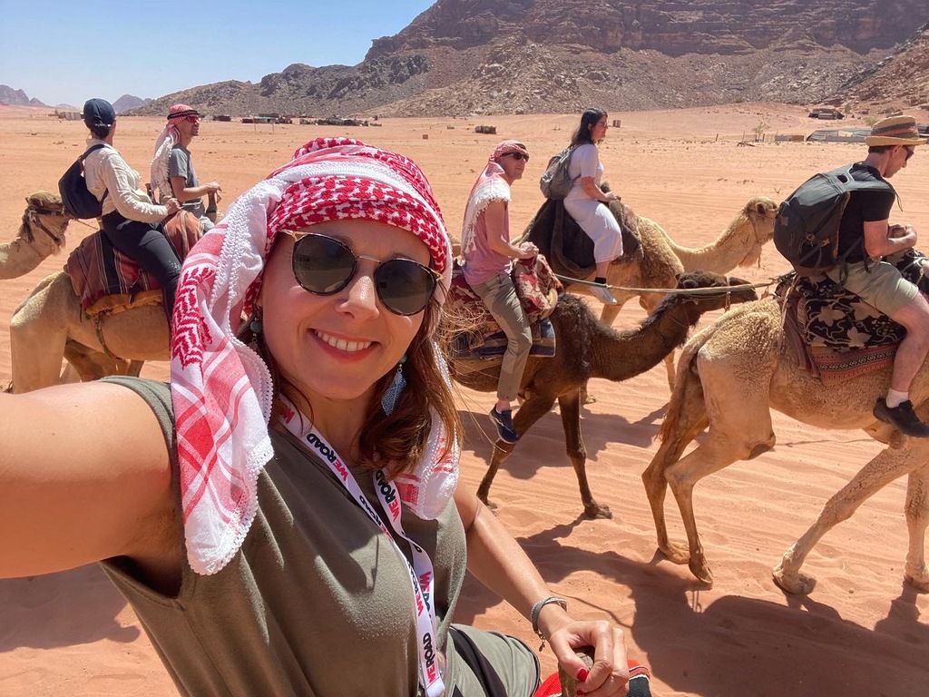 Une femme souriante portant un keffieh prend un selfie à dos de chameau, entourée de son groupe WeRoad lors d’une aventure dans le désert.