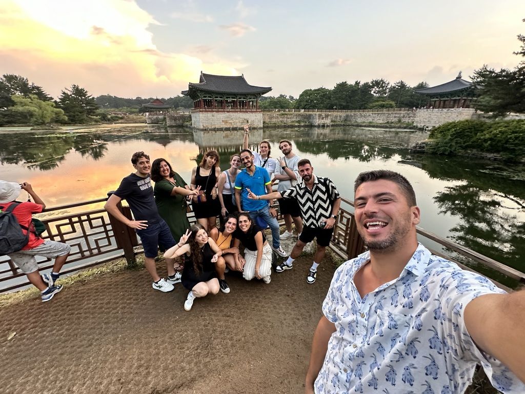 Un groupe de voyageurs WeRoad posant pour une photo devant un temple coréen traditionnel au coucher du soleil, entouré de nature et d’un lac.