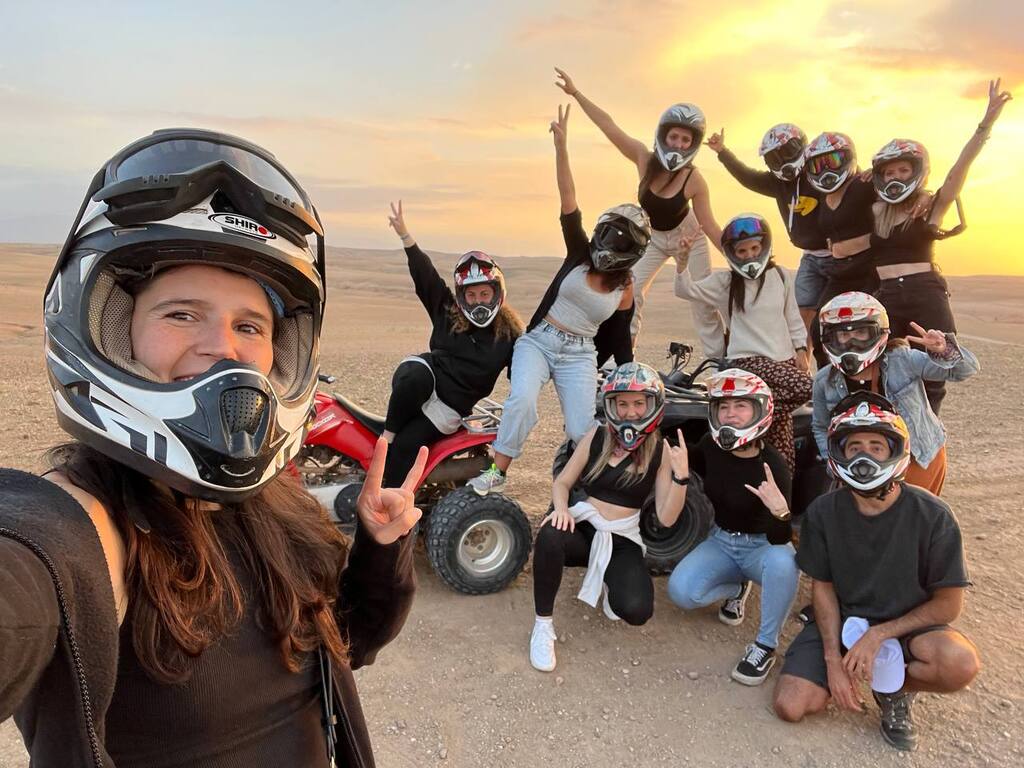 Groupe de voyageurs WeRoad en casque posant avec des quads sur le sable au coucher du soleil, dans une aventure pleine d’adrénaline.