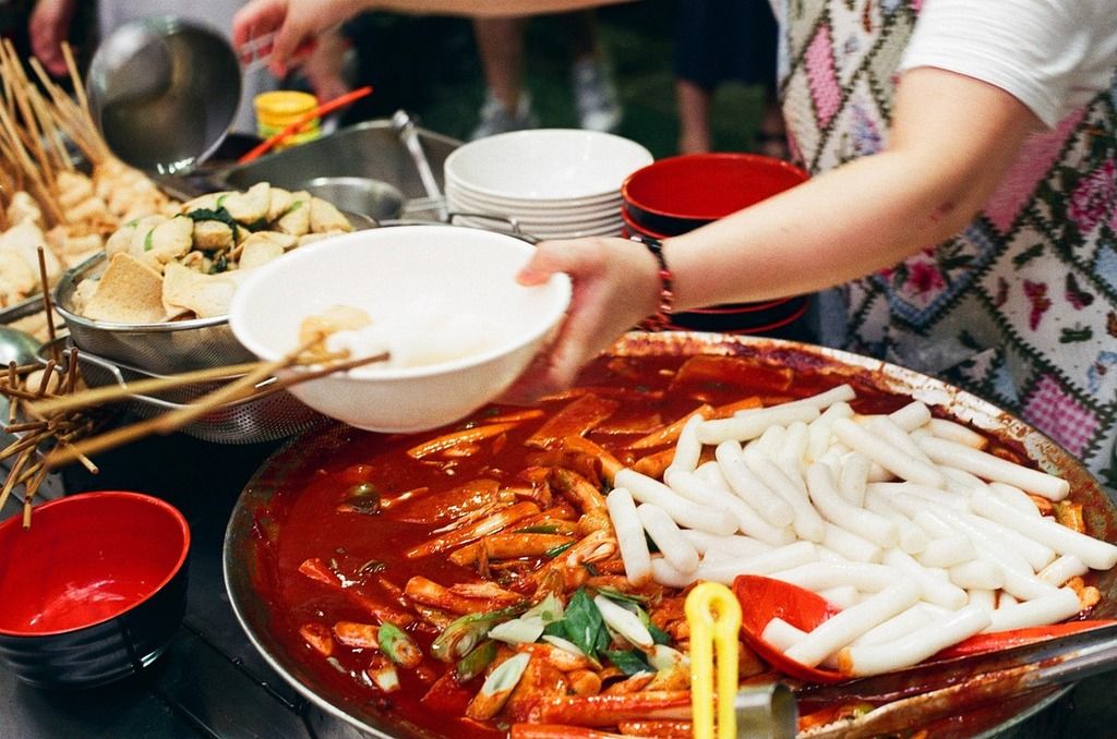 Un stand de street food coréenne servant du tteokbokki épicé avec des brochettes de poisson, des légumes et une sauce rouge pimentée.