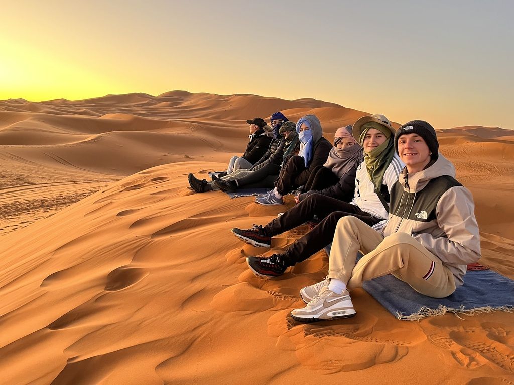 Groupe de voyageurs WeRoad assis sur les dunes du désert, admirant le lever du soleil dans une ambiance magique.