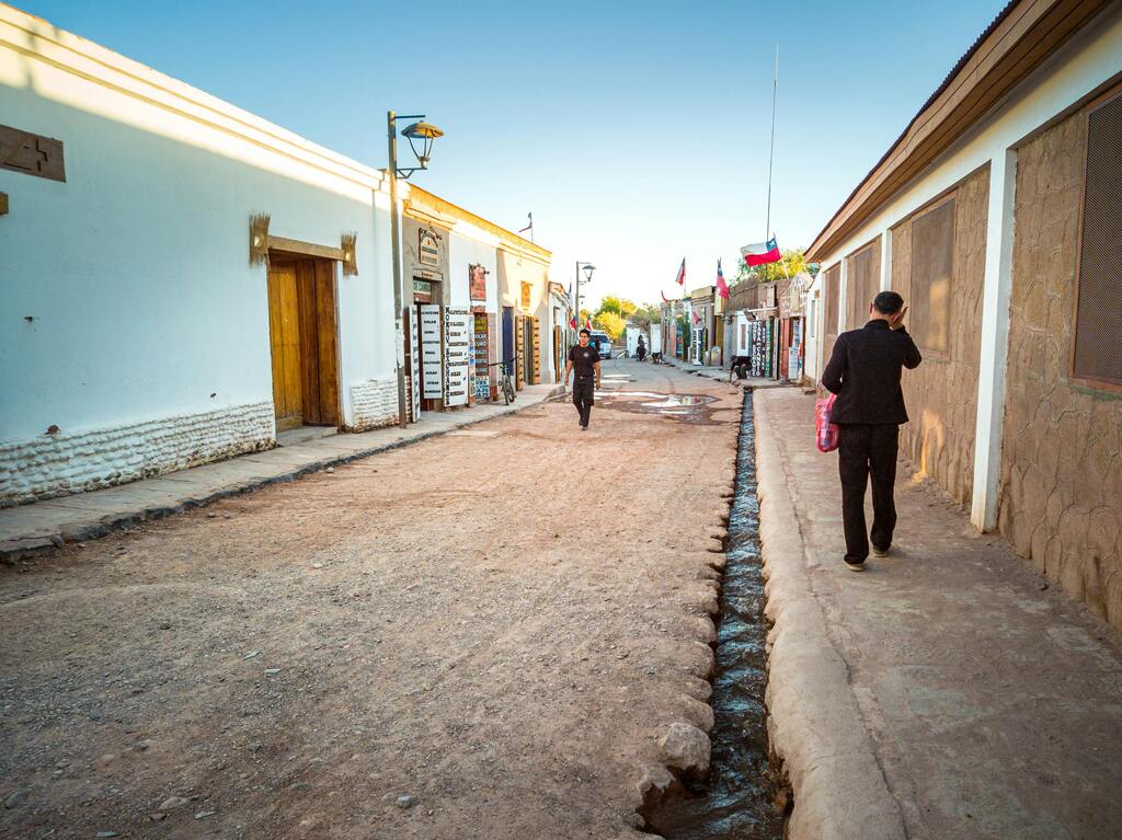 Rue de San Pedro de Atacama, Chili, avec des maisons traditionnelles en adobe et des habitants marchant sous un ciel bleu clair.