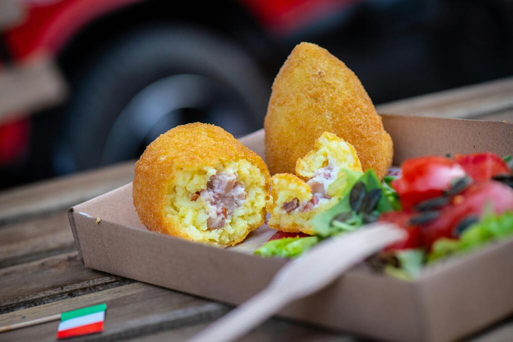 Deux arancinis siciliens dorés, l'un ouvert montrant une farce crémeuse au jambon, servis avec une salade fraîche dans une boîte en carton.