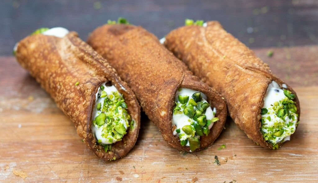 Trois cannoli siciliens croquants garnis de ricotta sucrée et de pistaches concassées sur une planche en bois.