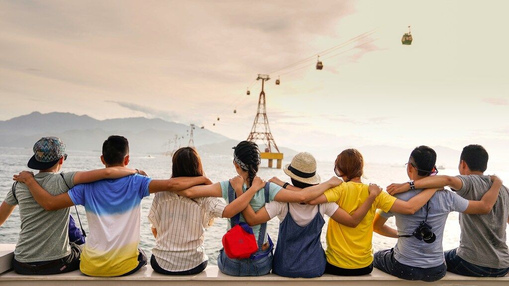 Groupe d’amis assis en ligne, les bras autour des épaules, contemplant la mer et les téléphériques à l’horizon.