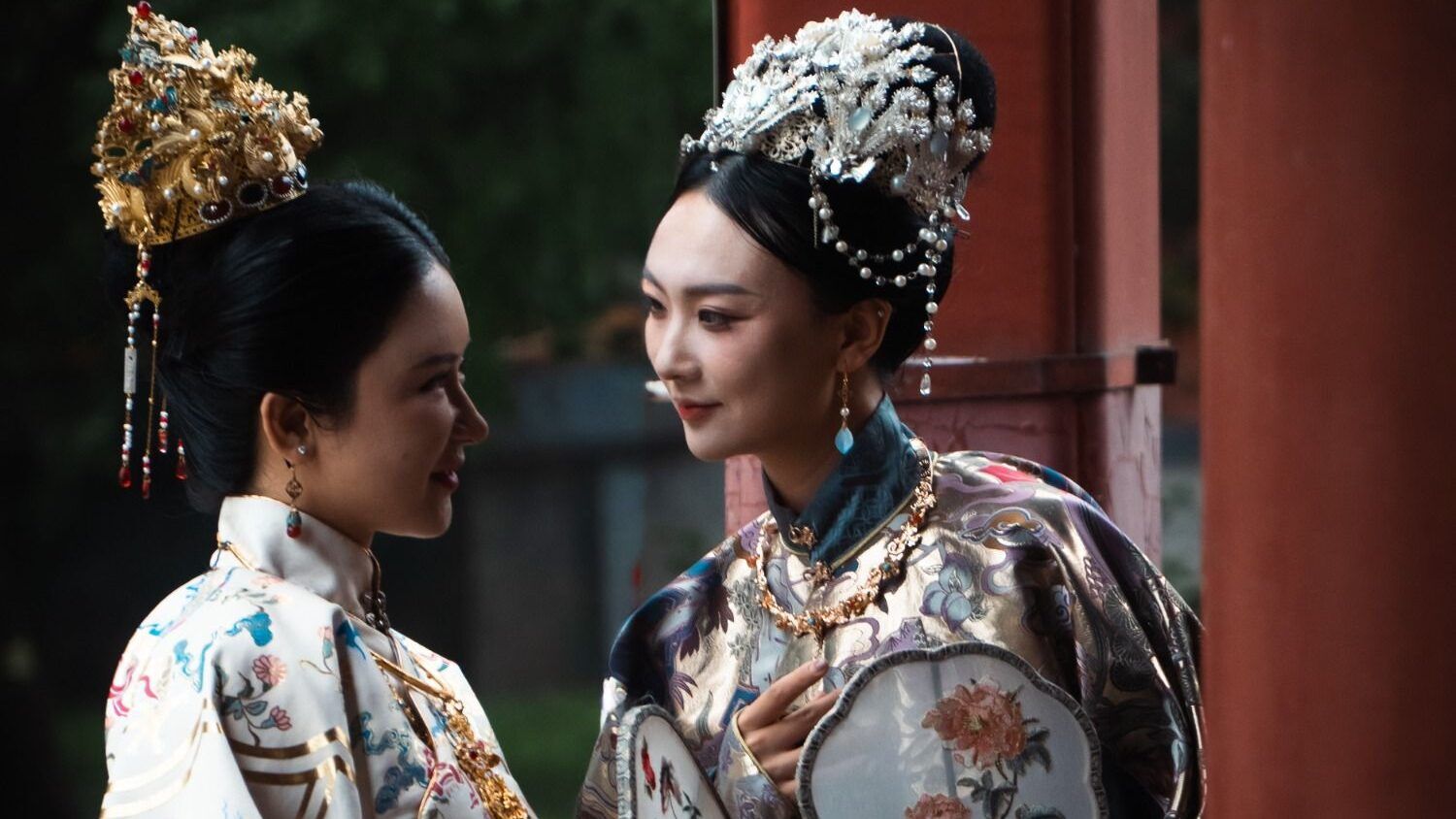 Deux femmes en costumes impériaux chinois et coiffures ornées, tenant des éventails ronds.