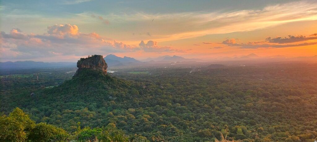 Vue panoramique sur le Rocher du Lion à Sigiriya, Sri Lanka, dominant une forêt luxuriante sous un ciel aux teintes orangées et rosées du coucher de soleil.