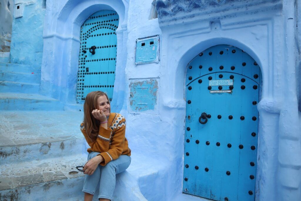 Jeune femme assise sur des escaliers bleus devant des portes marocaines traditionnelles à Chefchaouen.