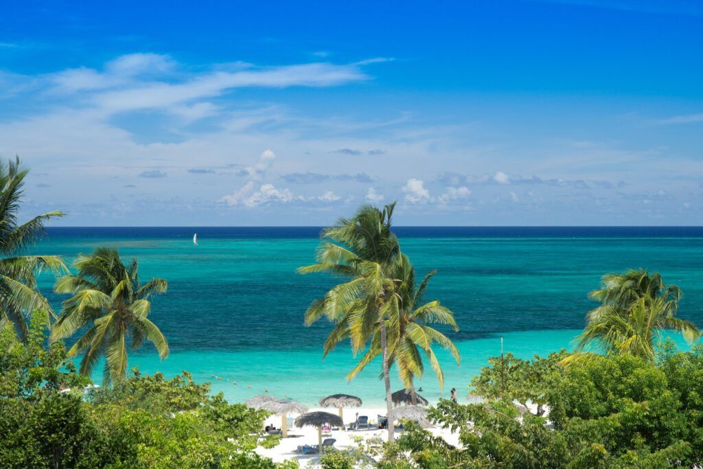 Vue en hauteur de la plage de Guardalavaca : palmiers, sable blanc et parasols de paille au bord d'une mer turquoise et bleue intense.