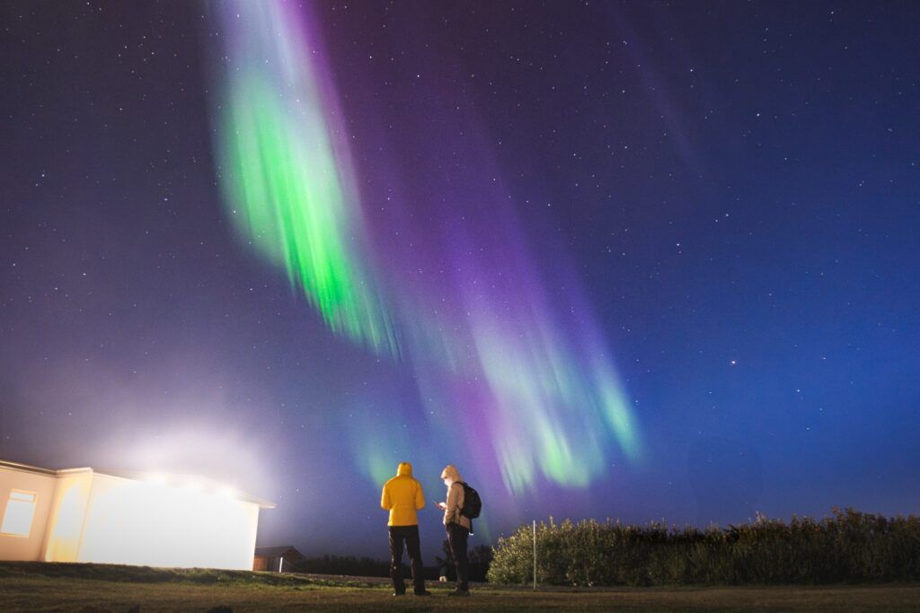 Two people contemplate a green and purple aurora borealis in a starry sky in Iceland.