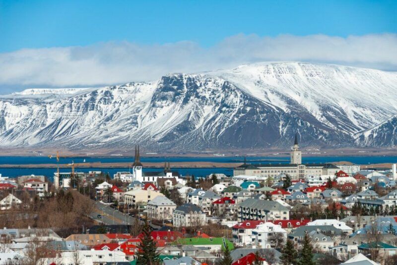Vue panoramique de la ville de Reykjavik avec ses maisons aux toits colorés, située au bord de l'eau devant une imposante montagne enneigée.