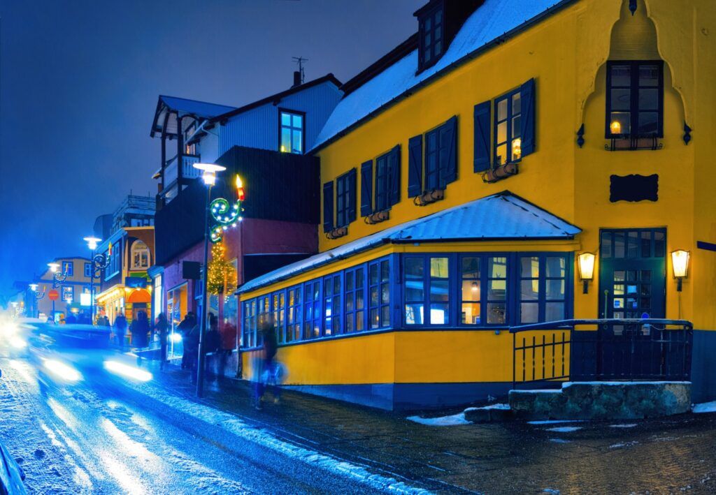 Rue Laugavegur enneigée de Reykjavik à la tombée de la nuit, mettant en avant une maison jaune vive illuminée avec des fenêtres bleues sous un ciel d'hiver.