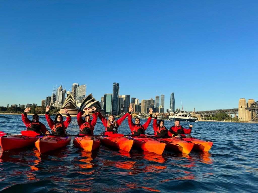 Un groupe de voyageurs WeRoad en kayak rouge saluant devant l'Opéra de Sydney sous un ciel bleu dégagé en Australie.