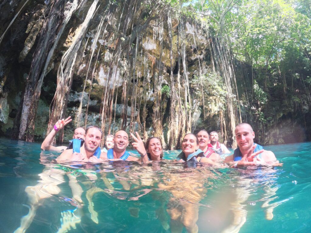 Un groupe joyeux de voyageurs WeRoad se baignant dans les eaux turquoise cristallines d'un cénoté au Mexique, avec d'impressionnantes racines d'arbres descendant le long des parois rocheuses en arrière-plan.